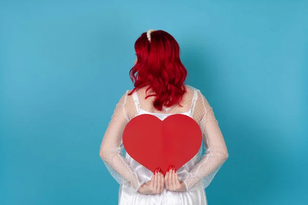 흰색 드레스를 입고 빨간 머리를 한 젊은 여성이 파란 배경에 고립 된 채 얼굴을 숨기고 있는 등 뒤에 빨간 종이 심장을 들고 있다 — 스톡 사진