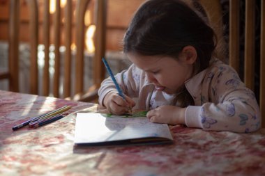 Hevesli bir kız renkli kalemlerle dilini uzatır ve ahşap bir evin oturma odasındaki masada çocuk dergilerine resim çizer.