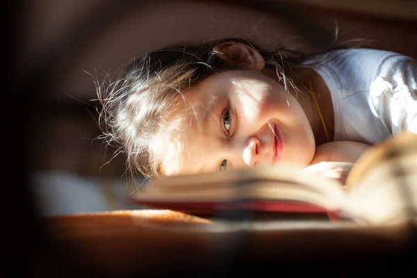 Retrato close-up através das barras de uma cama de ferro fundido de uma menina sorridente deitada de frente para baixo em um livro aberto à luz do sol do quarto — Fotografia de Stock