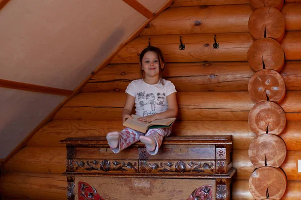 Egy csintalan, játékos lány ül egy öreg, kopott szekrényen feltartott lábakkal és egy nyitott könyvvel az ölében egy rusztikus faház hálószobájában. — Stock Fotó