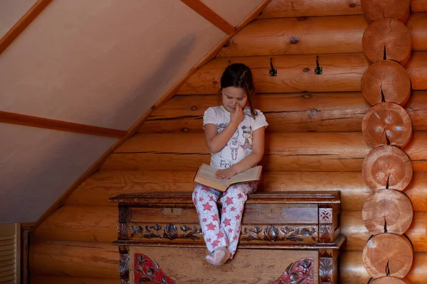 Zainteresowana dziewczyna siedzi na starym drewnianym kredensie, czytając książkę i drapiąc lub dłubiąc w nosie w sypialni rustykalnego domu z drewna — Zdjęcie stockowe