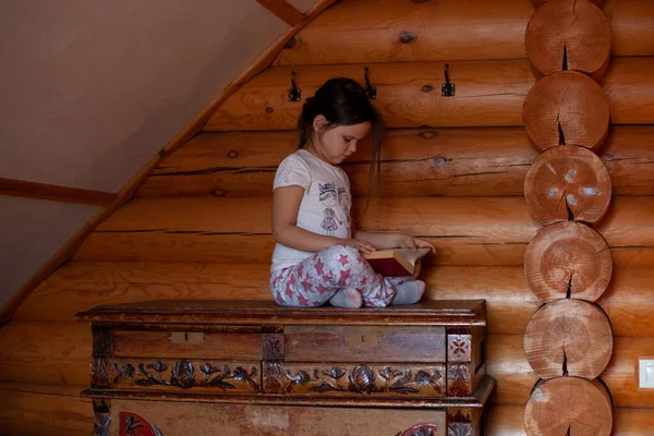 Egy figyelmes, komoly lány keresztbe tett lábbal ül egy régi, fából készült fiókos ládán, és könyvet olvas egy rusztikus faház hálószobájában. — Stock Fotó