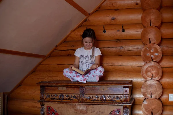 Przebiegła, uśmiechnięta dziewczyna siedzi ze skrzyżowanymi nogami na starym drewnianym kredensie i czyta książkę w sypialni rustykalnego domu z drewna — Zdjęcie stockowe