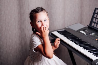 Beyaz yazlık elbiseli, ağzı kapalı, şaşırmış bir kızın yakın plan portresi ve bir evin içinde elektronik sentezleyicinin önünde oturan iki örgülü kız.