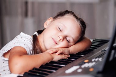 Beyaz bir elbise içinde gözleri kapalı ve iki örgülü saçı evde elektronik bir sentezleyicinin anahtarında uzanmış uyuyan bir kızın yakın plan portresi.