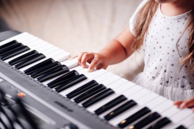 Yakın plan portre, uzun saçlı, beyaz elbiseli bir kızın eli oturma odasındaki elektronik bir sentezleyicinin tuşlarında melodi çalıyor.