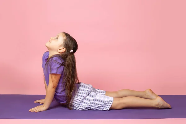 Full-length portrait of a girl in profile doing yoga and bhujangasana or upward dog pose, Cobra Pose on a purple mat isolated on a pink background — Stock Photo, Image