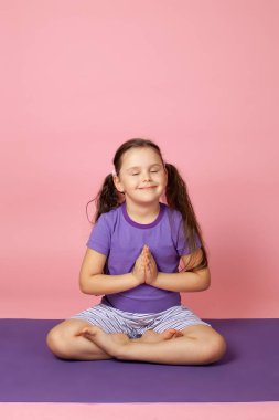 Huzurlu, mutlu, altı yaşında bir kız çocuğu yoga yapıyor, lotus pozisyonunda oturuyor ya da dua ediyor, pembe bir arka planda tek başına.