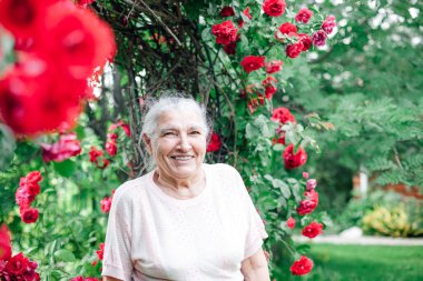 Gri saçlı yaşlı bir kadının yakın plan portresi. Geniş bir gülümsemesi ve gül çalılarının yanındaki bahçede kaplamaları var.