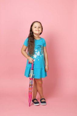  Mavi yazlık elbisesi içinde uzun dalgalı saçları olan güzel bir kız çapraz bacaklı ve elinde pembe bir arka planda izole edilmiş renkli bir şemsiye kamışı var.