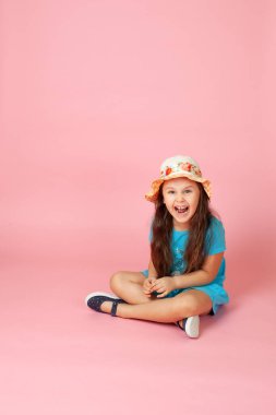 Kahkaha atan, uzun dalgalı saçlı, saman şapkalı, mavi elbiseli, yerde bağdaş kurmuş mutlu bir kızın portresi.