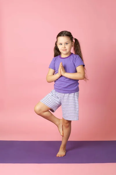 full-length portrait of a girl in shorts and a T-shirt on a purple mat in a tree pose in yoga, isolated on a pink background