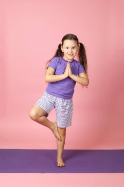 Beş yaşında şortlu bir kız çocuğu ve mor bir paspasın üzerinde tişört var. Ağaç duruşu yaparak yoga yapıyor, dengeyi geliştiriyor, pembe arka planda izole edilmiş.