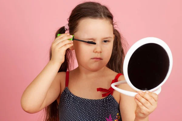portrait of a five-year-old girl painting her eyelashes with mascara and making up like her mother, isolated on a pink background