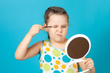 Beyaz elbiseli kızın yakın plan portresi yeşil göz farı sürüyor ve elinde mavi arka planda izole edilmiş beyaz plastik bir ayna tutuyor.
