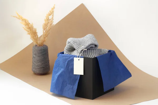 white blank paper logo tag on a grey knitted scarf in a black box and blue tissue paper and dried pampas grass in a spool yarn vase on beige paper