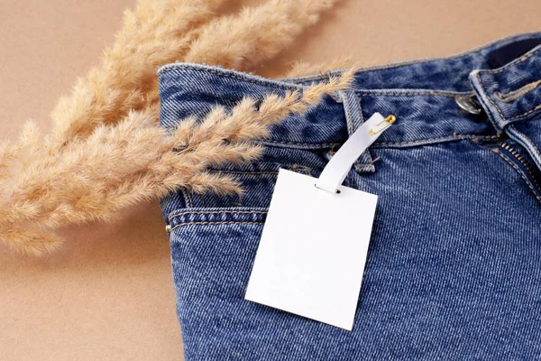 close-up white blank paper tag or label on a pin for a brand or logo on blue jeans with a decor of dry pampas grass or reeds