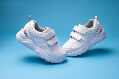 side view white kid sneakers with Velcro fasteners fly in the air in form of the letter V isolated on a blue background clipart