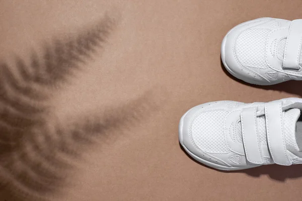 Επίπεδο θέσει δύο λευκά παιδικά sneakers με velcro συνδετήρες με φτέρη ή σκιά φοίνικα απομονώνονται σε ένα μπεζ φόντο χαρτί χειροτεχνίας — Φωτογραφία Αρχείου