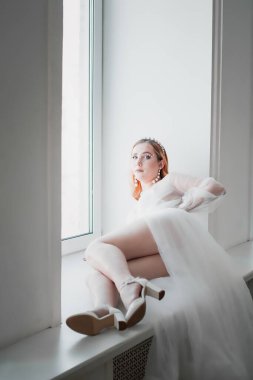 Gelin, beyaz şeffaf geceliğiyle, uzun bir trenle oturma odasındaki pencere eşiğinde uzanıyor.