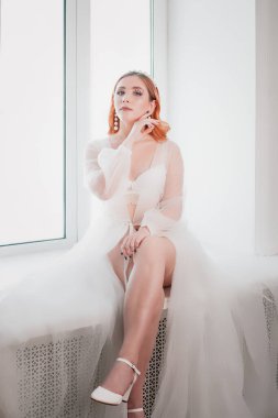 Beyaz şeffaf pelerinli, uzun boylu, genç bir gelin sabahleyin pencerede oturuyor.