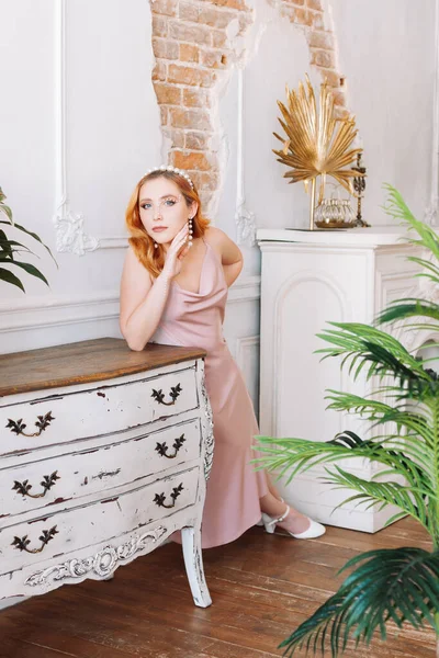 Een full-length portret van een roodharige vrouw in een beige zijden jurk leunend op haar elleboog op een oude ladekast in een studio met een armoedige bakstenen muur en huisplanten — Stockfoto