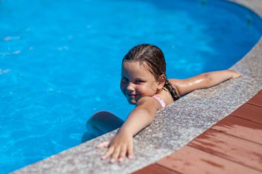 Gülümseyen beş yaşındaki bir kız ellerini otelin havuzunun kenarına koydu. Çocuklarla ailecek tatil yapma fikri.