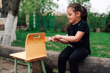 a five-year-old girl paints an old small chair with yellow paint for reuse while sitting on a log in the garden clipart
