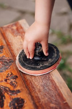 Bir çocuğun eli zımpara kağıdıyla bir rulo tutuyor ve bahçesinde çiçekli ahşap bir süs bulunan eski bir ahşap masayı temizliyor, geri dönüşüm ve sürdürülebilir.