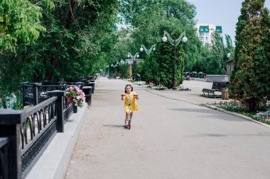 Bir kız şehir manzarasında sokak ışıkları, işlenmiş demir bir çit ve ağaçlarla motosiklet sürüyor.