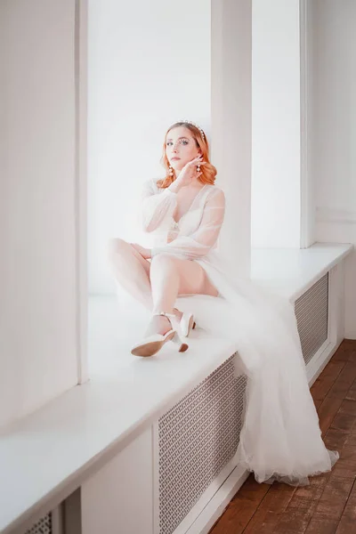 Een licht gevild noordelijk meisje in een witte transparante cape met een lange trein zit op de vensterbank in een lichte studio met grote ramen — Stockfoto