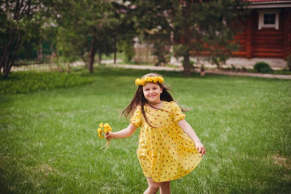 Una chica encantadora en un vestido amarillo baila con una corona de flores amarillas en su cabeza en un césped de hierba verde al aire libre en un día soleado de primavera — Foto de Stock