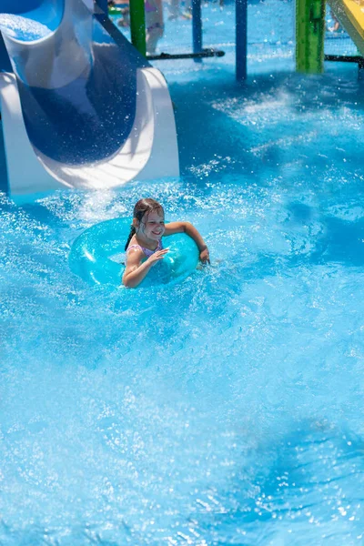 Una chica en un círculo inflable se desliza por un tobogán de agua en la piscina y envía salpicaduras de agua en todas las direcciones —  Fotos de Stock