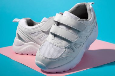 Geometrik kare kağıt üzerinde rahat ayakkabılar için cırt cırt cırtlı bağcıklı iki beyaz çocuk ayakkabısı, pembe ve mavi arka planda sert gölgeler.