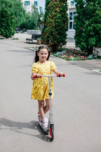 Pełnowymiarowy portret uroczej dziewczyny w żółtej letniej sukience jeżdżącej na skuterze po asfaltowej ścieżce wzdłuż alejki z cyprysami — Zdjęcie stockowe