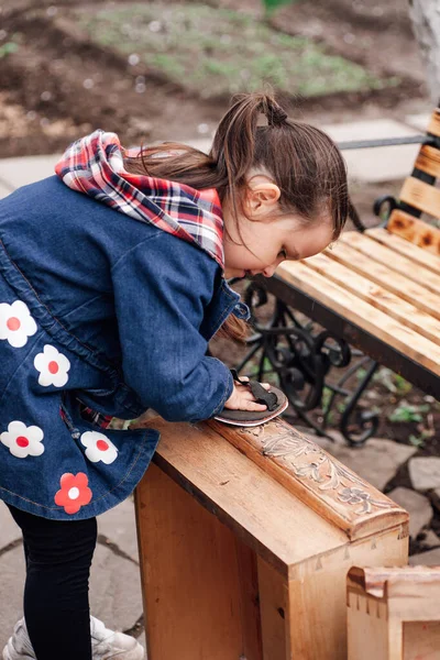 Una ragazza aiuta a carteggiare una vecchia cassettiera in legno con un rotolo di carta vetrata, riciclando mobili vintage — Foto Stock