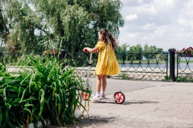 Sarı elbiseli bir kızın yeşil çiçek tarhlı bir yolda scooter sürüşünün arka görüntüsü. Bir kız yaz günü nehir boyunca scooter sürmeyi öğreniyor.