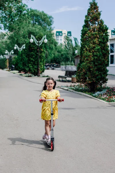 Una ragazza carina in un prendisole giallo impara a guidare uno scooter in giro per la città e gode di una calda giornata estiva — Foto Stock