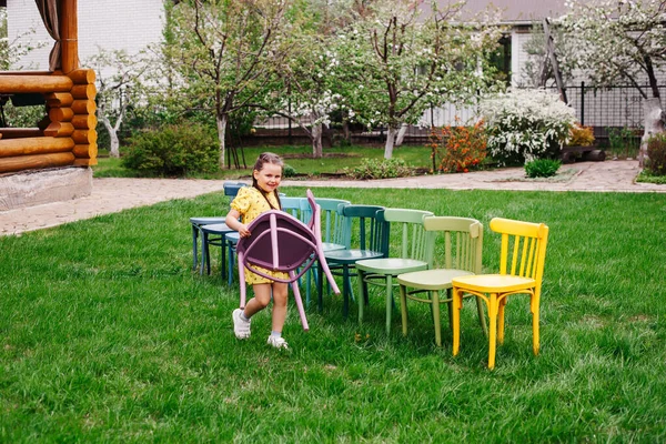 Egy sárga ruhás lány fából készült széket hord, és sorba rendezi a székeket, megszervezi a gyerekek születésnapi partiját a kertben nyáron. — Stock Fotó