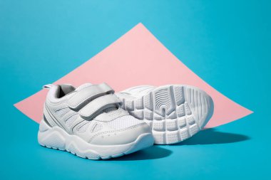 Makro kenarlı bir bakış açısı. Bir beyaz kız ayakkabısı ikinci bir spor ayakkabısının önünde duruyor. Tabanı geometrik kare bir kağıtta, pembe ve mavi arka planda kameraya bakıyor.