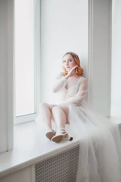 De bruid in een witte transparante negligé met een lange trein zit met haar voeten op de vensterbank — Stockfoto