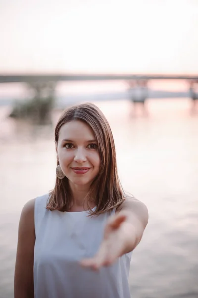 Una mujer caucásica sonriente feliz extiende su mano a la cámara y ofrece ir a una cita o en un viaje, un puente a través del río al atardecer en colores pastel —  Fotos de Stock