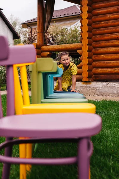 Egy vidám lány mászik végig egy sor színes fa székek, egy lány jól érzi magát a kertben — Stock Fotó