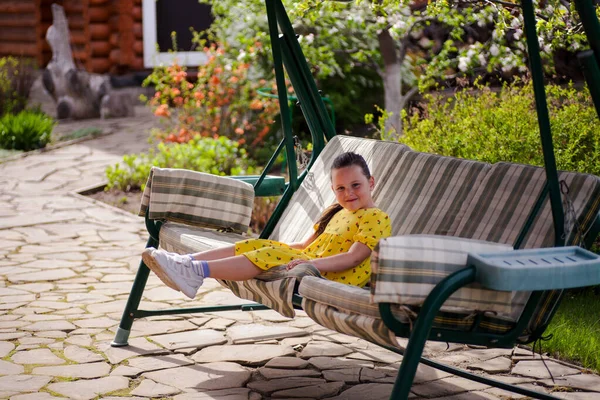 Egy gyönyörű, sárga ruhás lány ül a hintán a kertben, egy virágzó tavaszi kertben, egy fényes, napos napon, nyaralni a családjával. — Stock Fotó