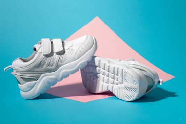 Makro kenarlı bir bakış açısı. Bir beyaz çocuk, ikinci bir koşu ayakkabısının tabanında durur. Geometrik kare bir kağıtta, pembe ve mavi arka planda yatar.