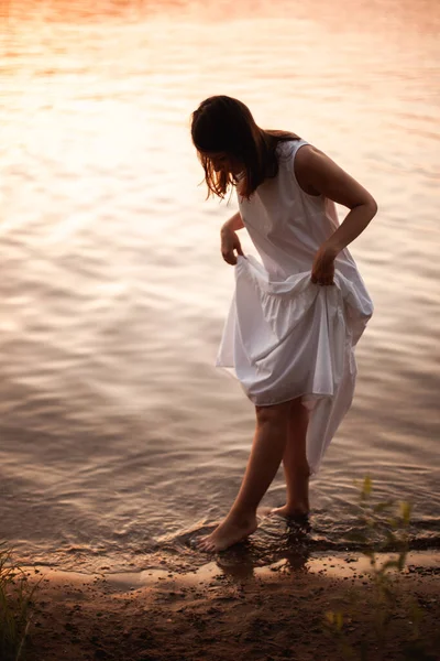 Die Silhouette einer jungen Frau in einem weißen Sommerkleid, die an einem warmen Sommerabend am Ufer des Flusses am Ufer des kühlen Wassers entlang geht, ein orangefarbener Sonnenuntergang und die orangefarbene Oberfläche des Flusses — Stockfoto