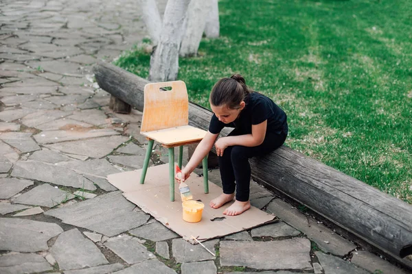 Portré egy teljes hosszúságú gyermekről, aki egy széket fest újra használatra, és egy fatörzsön ül egy köves úton a pázsit mellett egy nyári napon — Stock Fotó