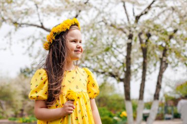 Kafasında sarı karahindiba çelengi ve elinde bahar gökyüzü ve arka planında ağaçlar olan bir buket karahindiba olan gülümseyen bir kızın portresi.