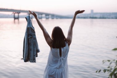 Yaz yaşam tarzı, beyaz elbiseli genç bir kadın mutlu bir şekilde ellerini sallıyor ve gün batımında şehrin siluetini seyrederek iyi vakit geçiriyor.