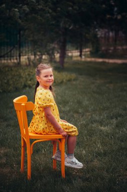 Sarı elbiseli sevimli bir kızın portresi. Çimenlikteki turuncu sandalyede oturuyor. Yaz günbatımında bir parkta.